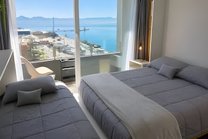 A hotel room with a large bed, a smaller bed, and a balcony with a view of the sea and mountains.