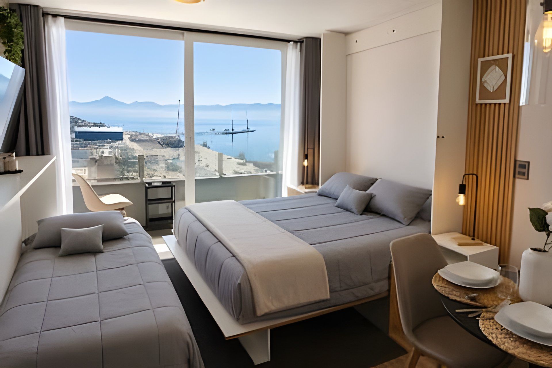A hotel room with two beds, a desk, and a view of the sea and mountains.