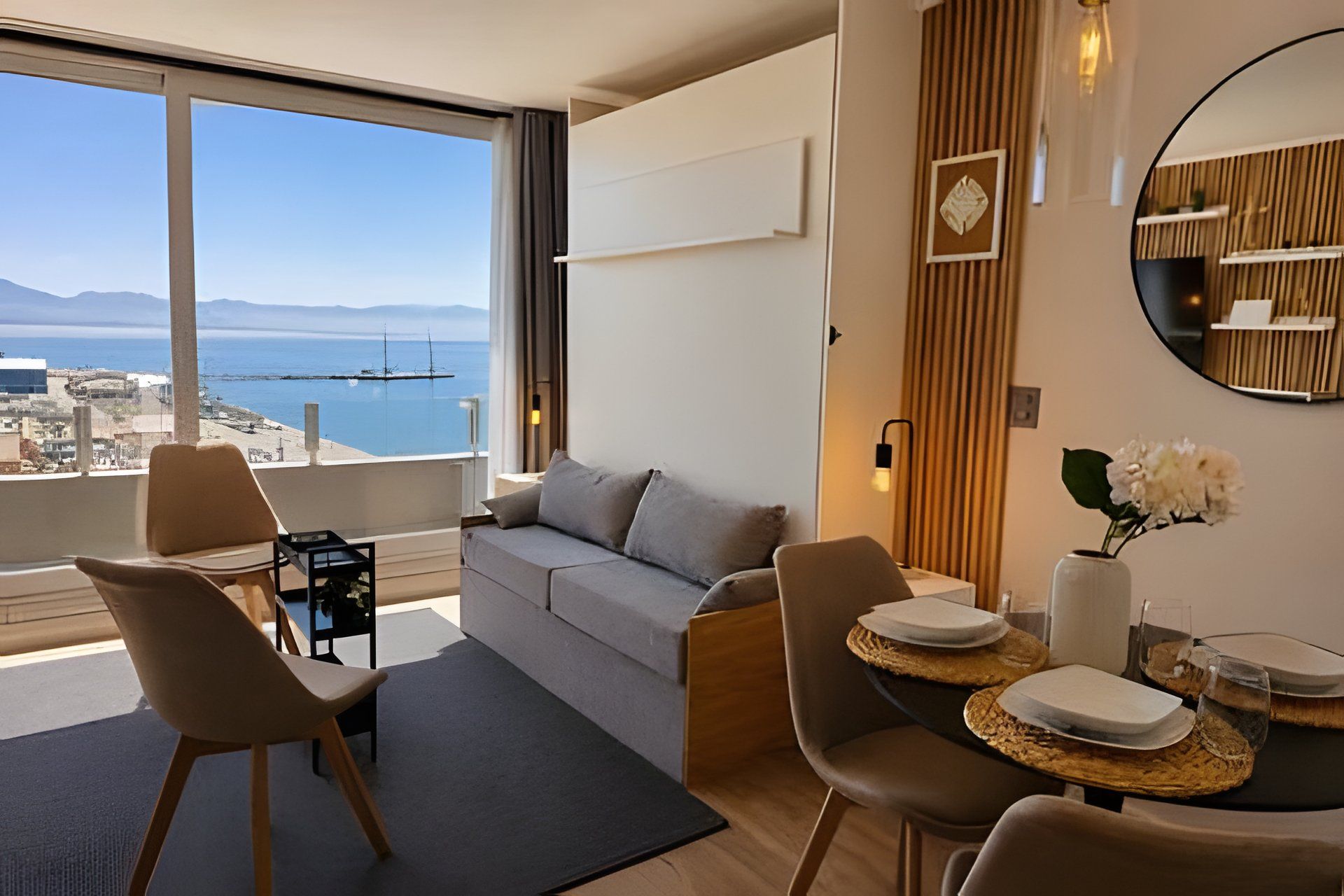 A modern living room with a sofa, chairs, a dining table with plates, a vase with flowers, and a large window with a view of the sea and buildings.