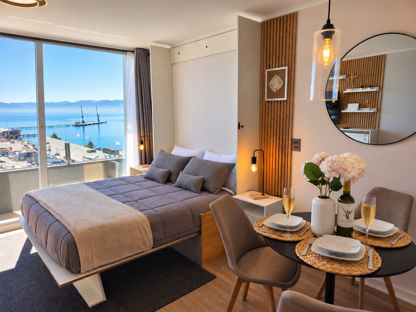 Una habitación de hotel moderna con cama, un área de comedor con una mesa para dos personas y un amplio ventanal con vistas al mar y un muelle.