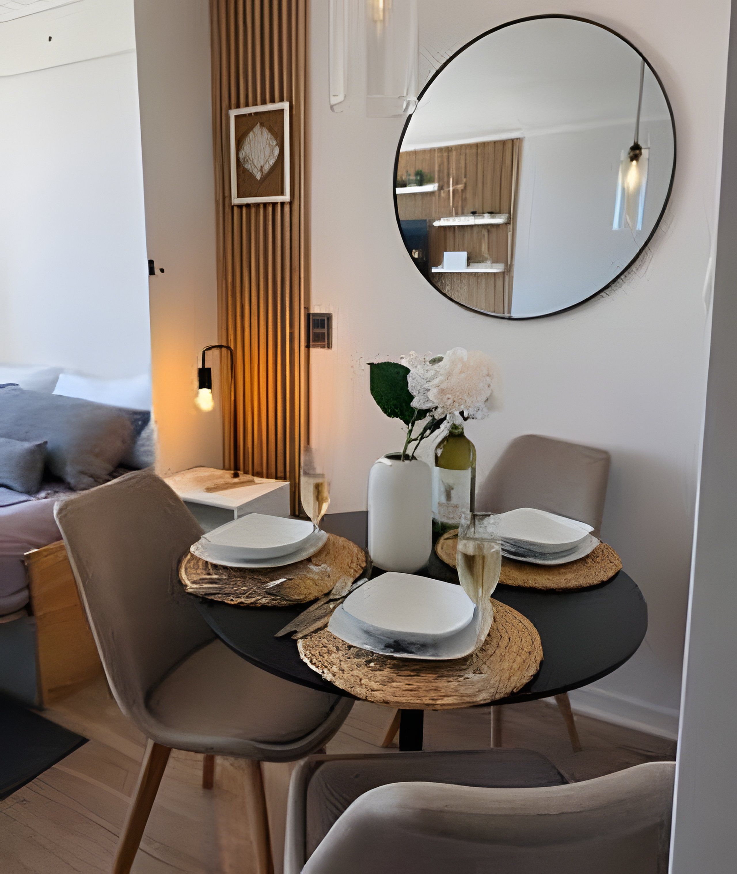 A dining area with a round table, two chairs, a vase with flowers, and a mirror on the wall.