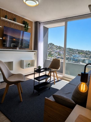 A modern living room with a large window, a flat-screen TV, and a view of a hillside town.