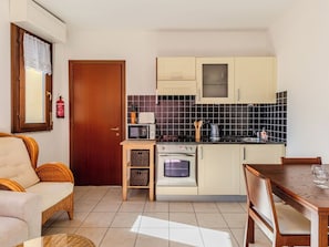 Cocina compacta con microondas, tostadora, hervidor y horno. Hay una mesa de comedor con sillas, un sofá de mimbre y una pequeña mesa de madera con vasos.