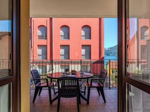 Ein Balkon mit rundem Tisch und Stühlen, Blick auf ein rotes Gebäude mit Rundbogenfenstern und ein Gewässer im Hintergrund.
