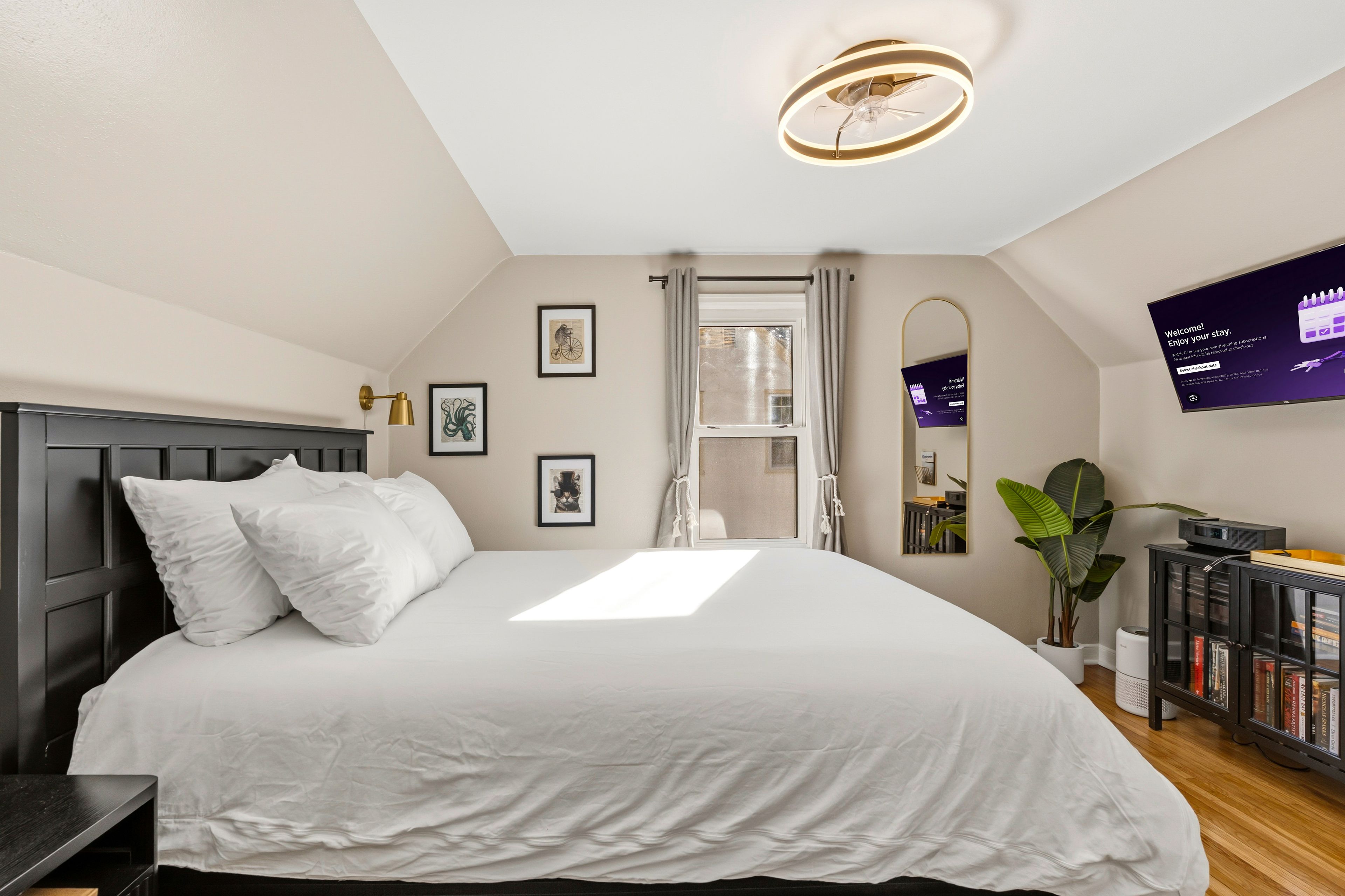 A bedroom with a large bed, a ceiling light, a window with curtains, a wall-mounted TV, and a bookshelf.