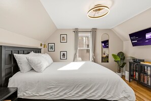 Un dormitorio con una cama grande, una lámpara en el techo, una ventana con cortinas, un televisor montado en la pared y una estantería.