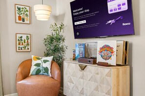 A modern living room with a leather armchair, a wall-mounted TV, framed botanical prints, and a wooden console with books and a record player. - Mayo Clinic Cottage - Power Bed & Recliners (Rochester)