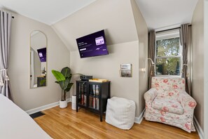 A modern living room with a flat-screen TV, a bookshelf, a potted plant, a floral armchair, and a window with curtains. - Mayo Clinic Cottage - Power Bed & Recliners (Rochester)