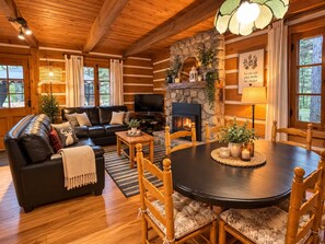 A cozy log cabin interior with a fireplace, a black leather sofa, a dining table, and wooden chairs. - Le Shack Shack | Chalet en bois • Spa • Accès au lac (Val-des-Lacs)