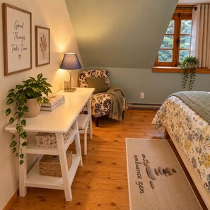 A cozy bedroom with a wooden floor, a bed with floral bedding, a desk with a lamp, a potted plant, and a window with curtains. - Le Shack Shack | Chalet en bois • Spa • Accès au lac (Val-des-Lacs)