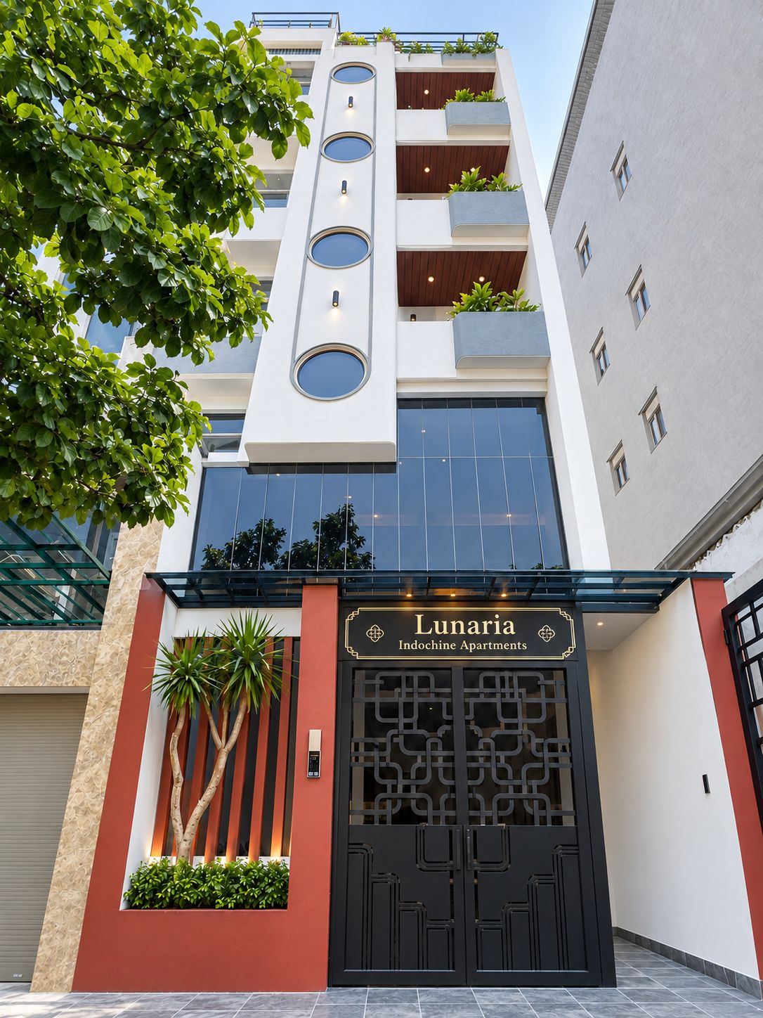 Un edificio moderno con una amplia entrada negra, balcones con plantas y un letrero que dice 'Lunaria Indochine Apartments'.