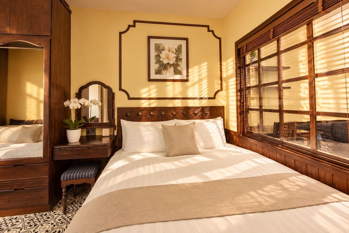 A hotel room with a large bed, a wooden wardrobe, a bedside table with a mirror, a potted plant, and a window with blinds.