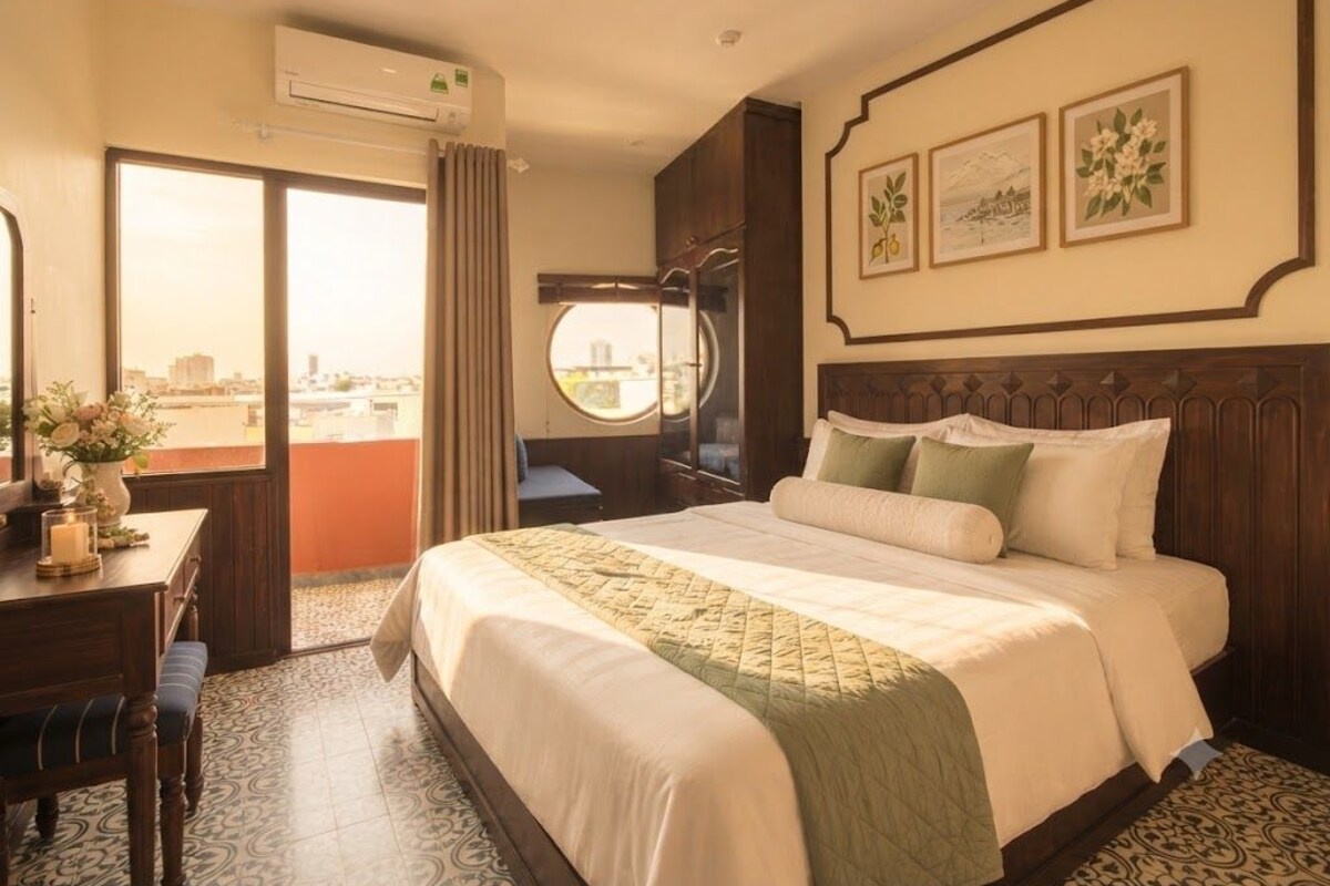 A hotel room with a large bed, a desk with a chair, a window with curtains, and a view of the city.