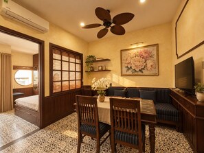 Une pièce comprenant une table à manger, des chaises, un canapé, un téléviseur, un ventilateur de plafond et une grande fenêtre avec des stores.