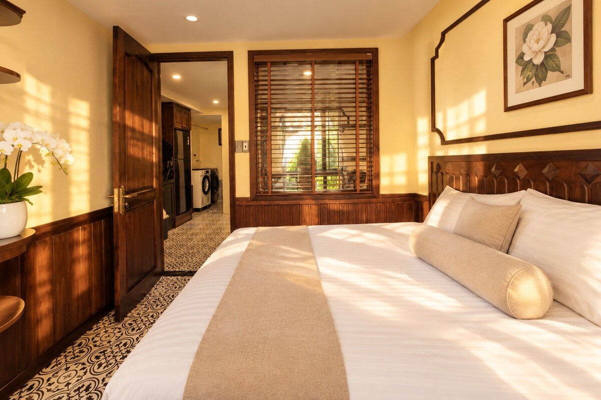 A hotel room with a large bed, a wooden headboard, a potted plant, and a view through a window with blinds.