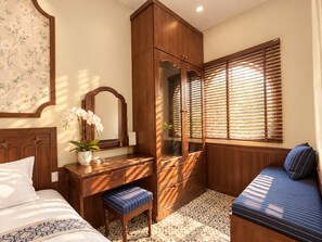 A bedroom with a bed, a wooden dresser, a mirror, a bench, and a window with blinds. - Lunaria Indochine Apartments (An Hai)