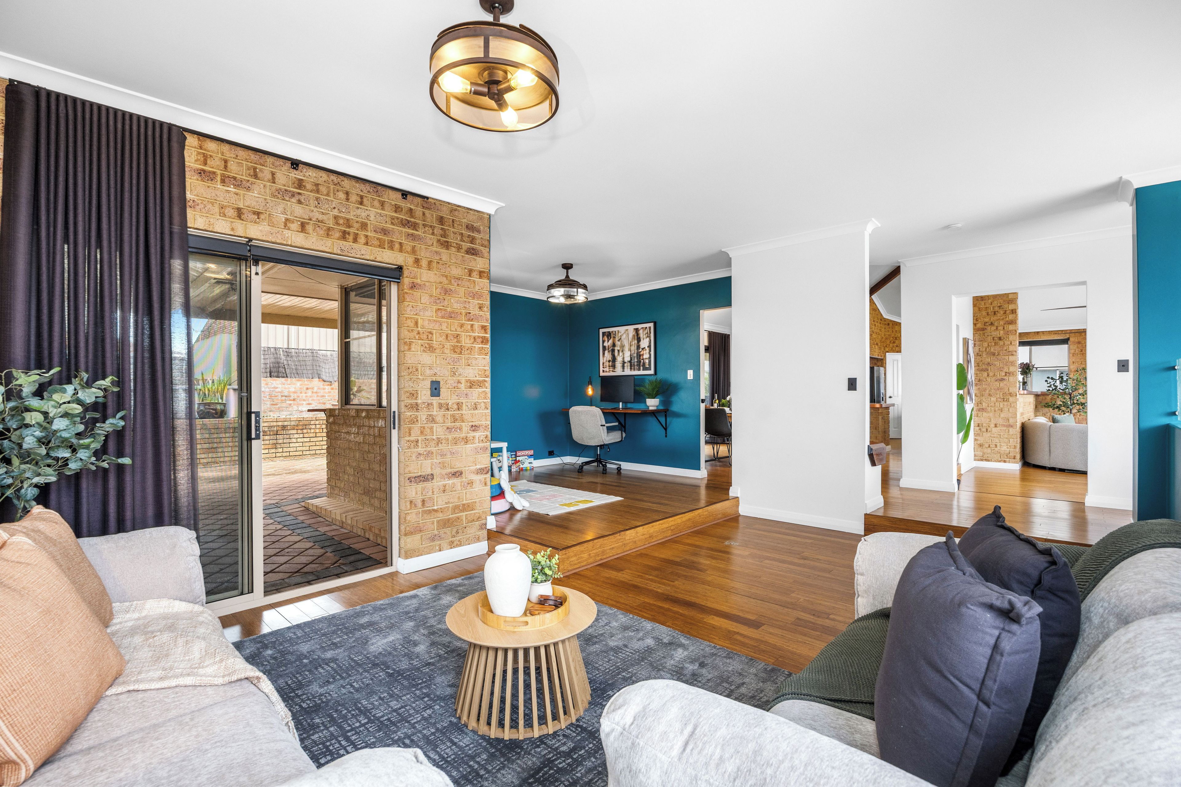A modern living room with a brick wall, a sliding glass door leading to a balcony, a dining area with a blue accent wall, and a kitchen in the background.