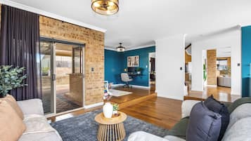 A modern living room with a brick wall, a sliding glass door leading to a balcony, a dining area with a blue accent wall, and a kitchen in the background.