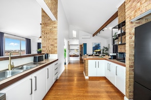 A modern kitchen with white cabinetry, a black refrigerator, and a view of the city through large windows. - The Beach House | 4BR, Spa & Jacuzzi near Coogee Beach (Coogee)