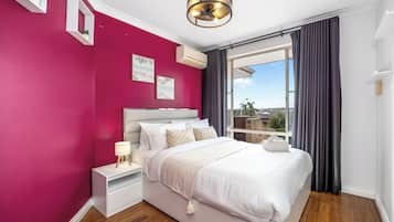 A modern bedroom with a large bed, a nightstand, a lamp, and a view of the outdoors through a window.