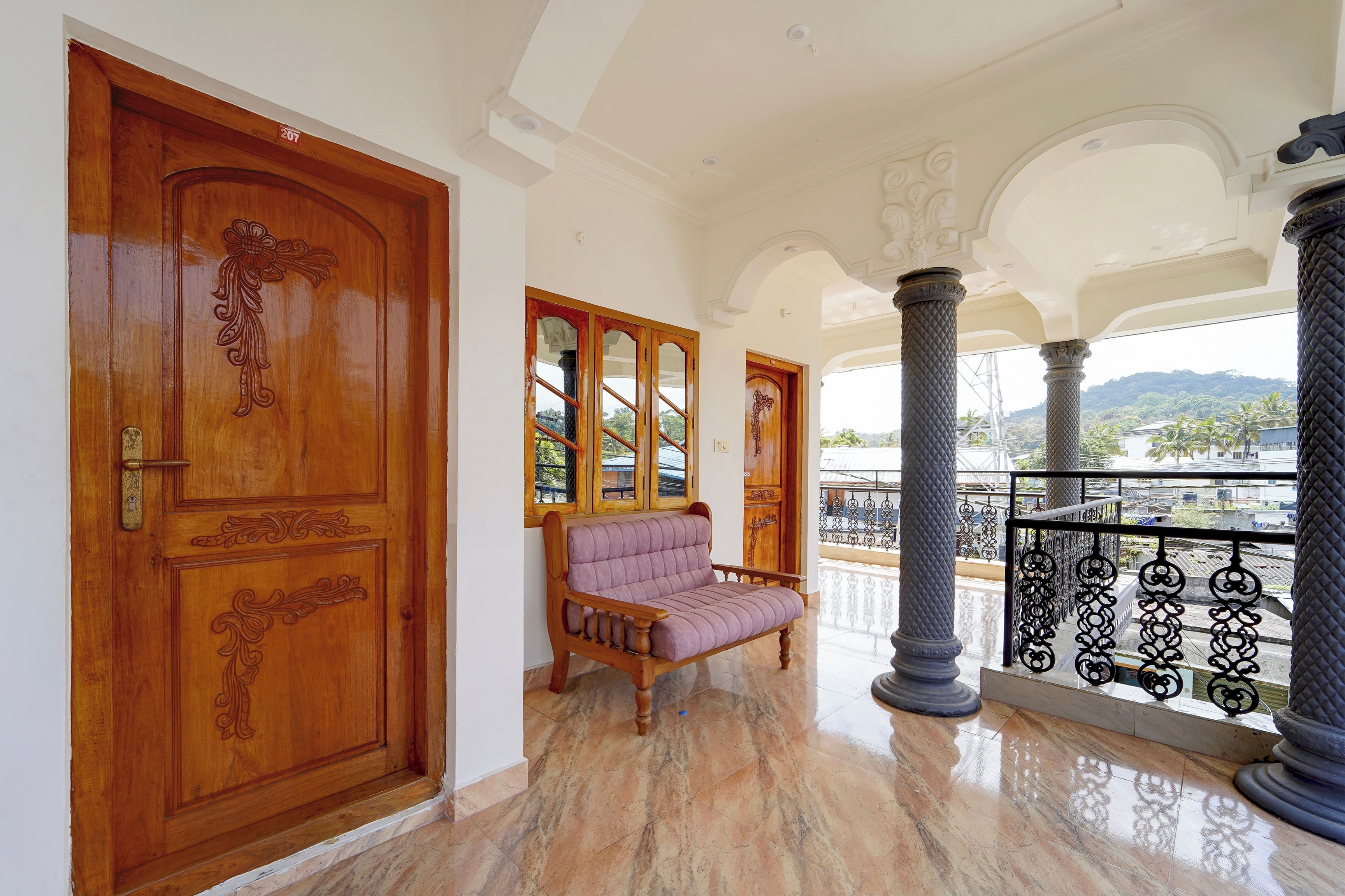 A spacious hallway with a wooden door, a cushioned bench, and a balcony with a metal railing.