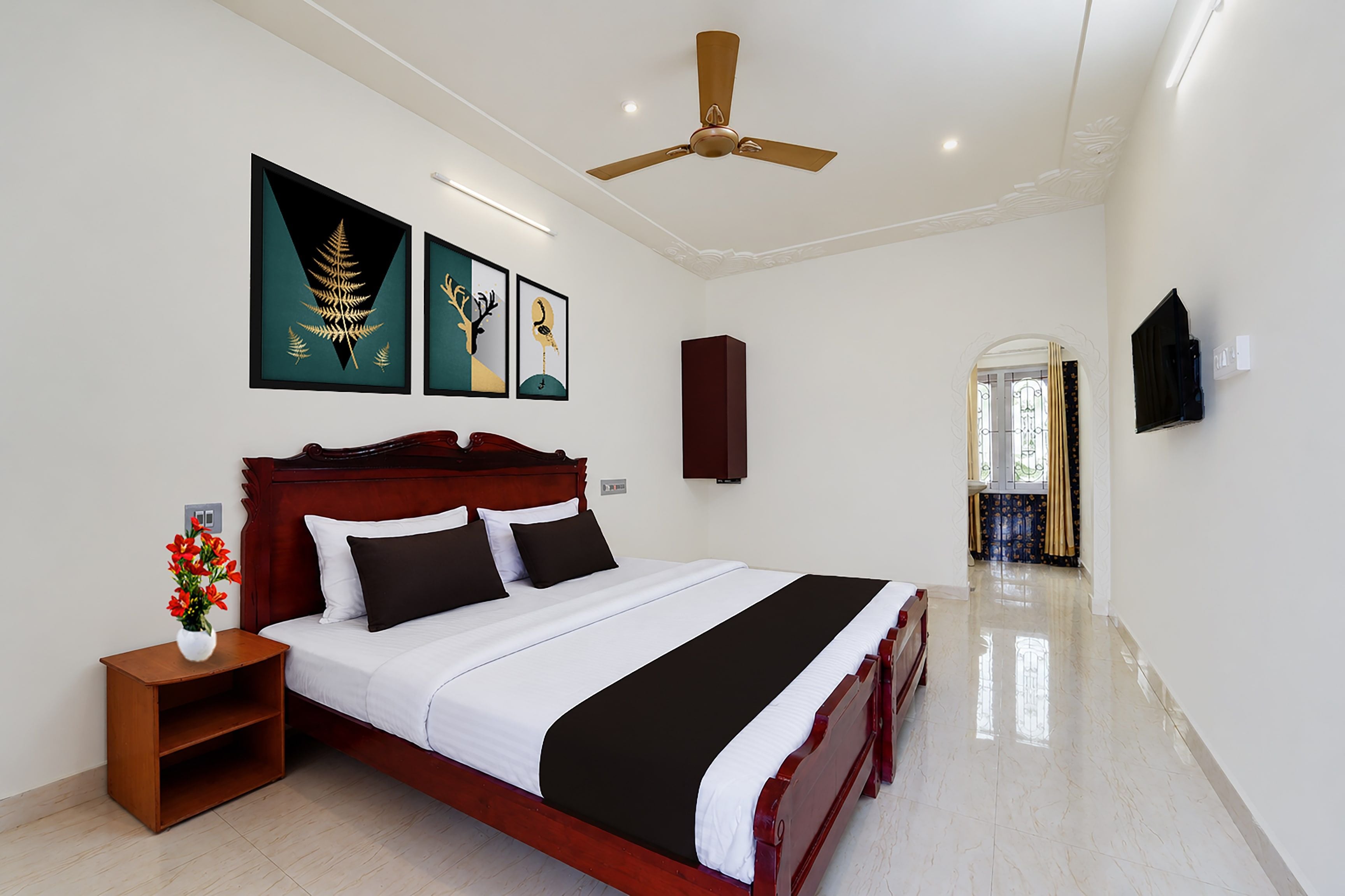 Quarto com cama, mesa de cabeceira, vaso com flores, ventoinha no teto e obras de arte na parede.
