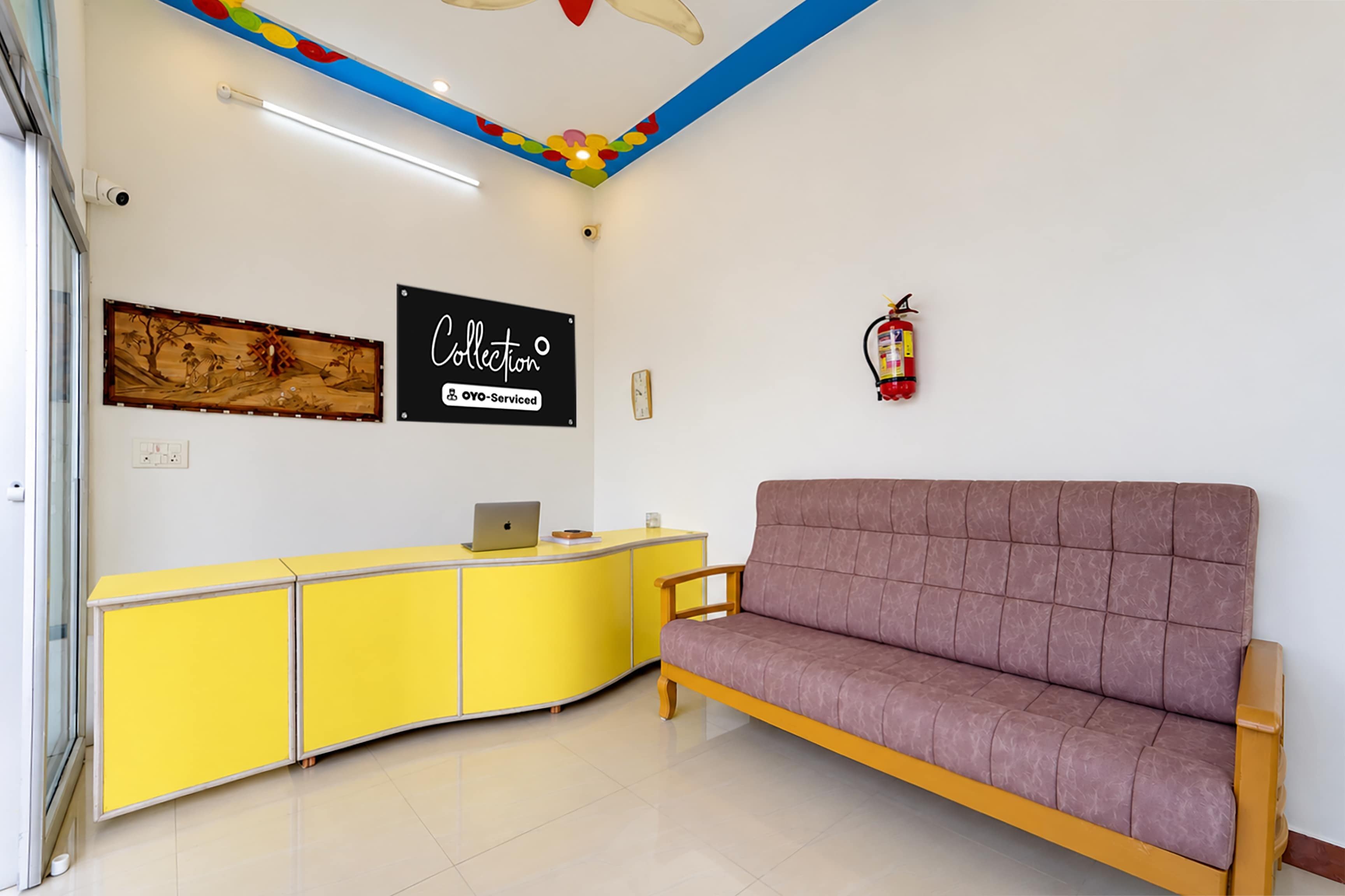 Uma área de recepção com uma mesa amarela, um sofá roxo, um extintor de incêndio e uma placa na parede com a palavra 'Collection'.