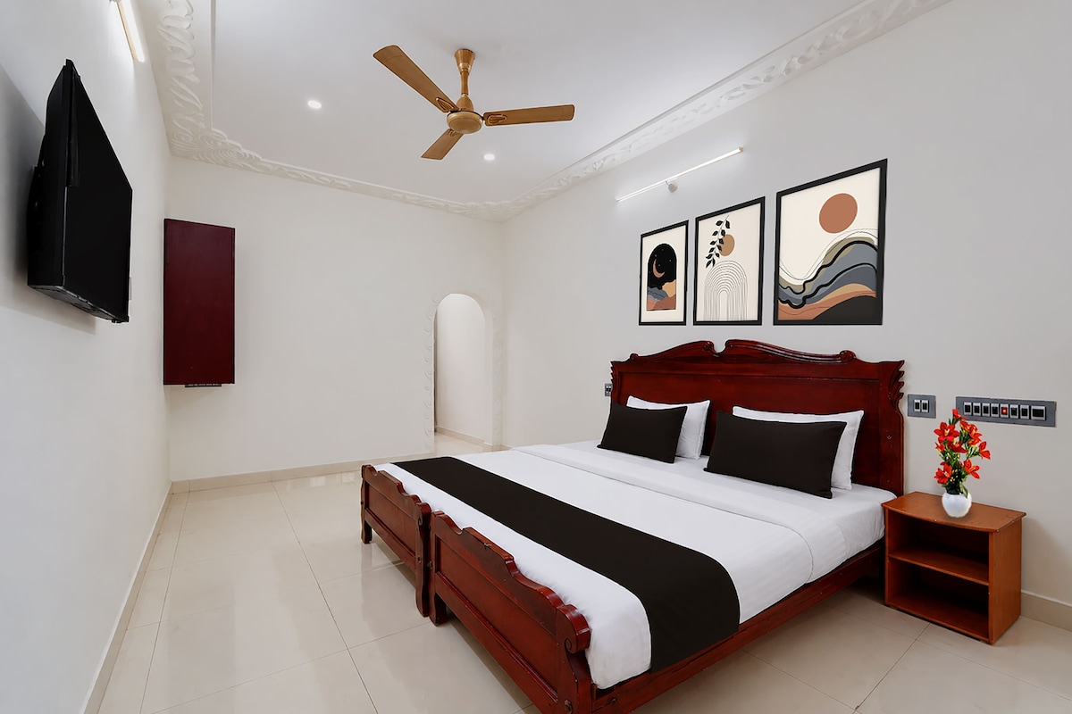 A bedroom with a wooden bed, a ceiling fan, a TV, artwork on the wall, and a nightstand with a vase of flowers.
