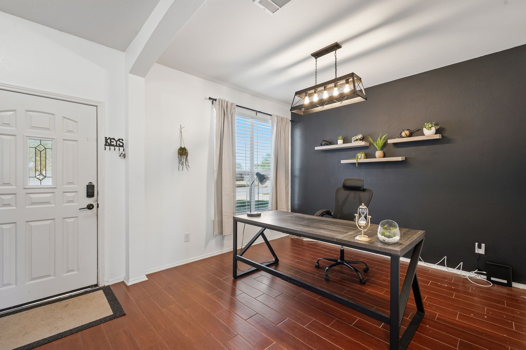 Un interior moderno con una mesa de comedor de madera, una silla de oficina negra, una lámpara colgante y una pared negra con estantes que sostienen plantas y objetos decorativos.