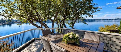 An outdoor deck with a wooden table and wicker chairs, overlooking a calm lake with trees and a clear sky.