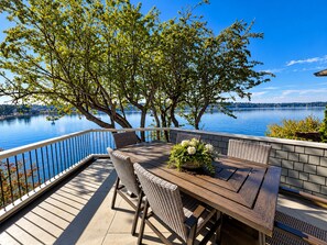 An outdoor deck with a wooden table and wicker chairs, overlooking a calm lake with trees and a clear sky. - Historic Puget Sound Waterfront • Capitol + Marina View • Walk to Farmers Market (Olympia)