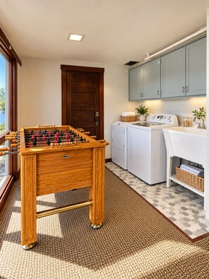 Een kamer met een houten tafelvoetbalspel, een wasmachine, een droger en een kast met een plant erop.