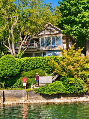 A house with a balcony overlooking a body of water, surrounded by lush greenery. - Historic Puget Sound Waterfront • Capitol + Marina View • Walk to Farmers Market (Olympia)