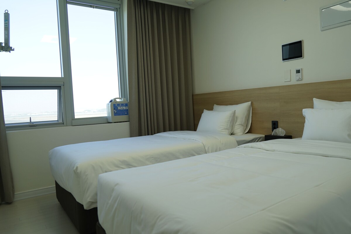 A hotel room with two beds, a window with curtains, a bedside table with a bottle, a digital thermostat, and a small screen on the wall.