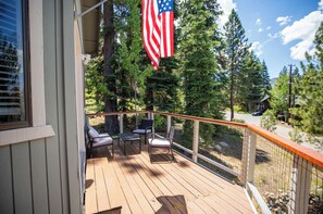 Una terraza de madera con mobiliario exterior, vista a árboles y una bandera estadounidense.