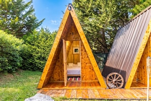 Ein dreieckiges A-Zelt aus Holz mit einer Liege im Inneren, einem kleinen Fenster und einem Rad an der Außenwand.