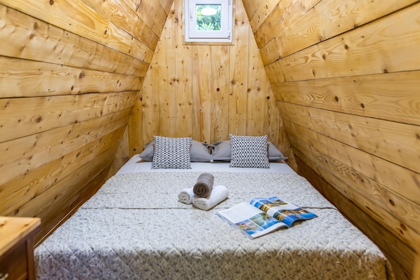 Ein kleiner, hölzerner Raum mit schräg stehender Decke, einem Bett mit zwei gemusterten Kissen, einem zusammengerollten Handtuch und einer Zeitschrift auf dem Bett.