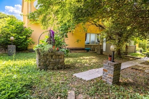 Ein Gartenbereich mit einem Steinpflanzkübel, einem Weg und einem Gebäude mit einer gelben Wand im Hintergrund.