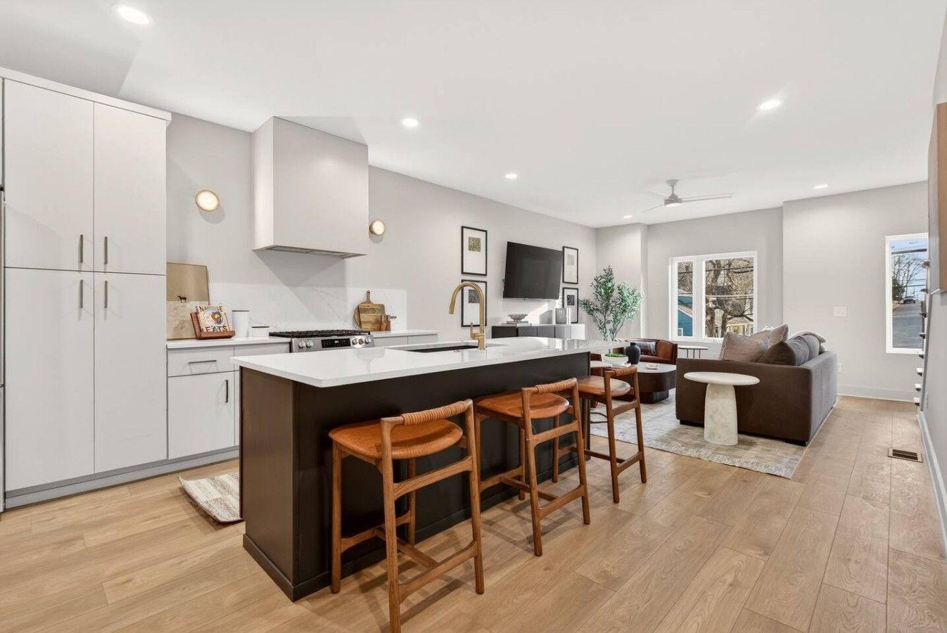 A modern kitchen with a central island, bar stools, a dining area with a sofa, and a large window providing a view.