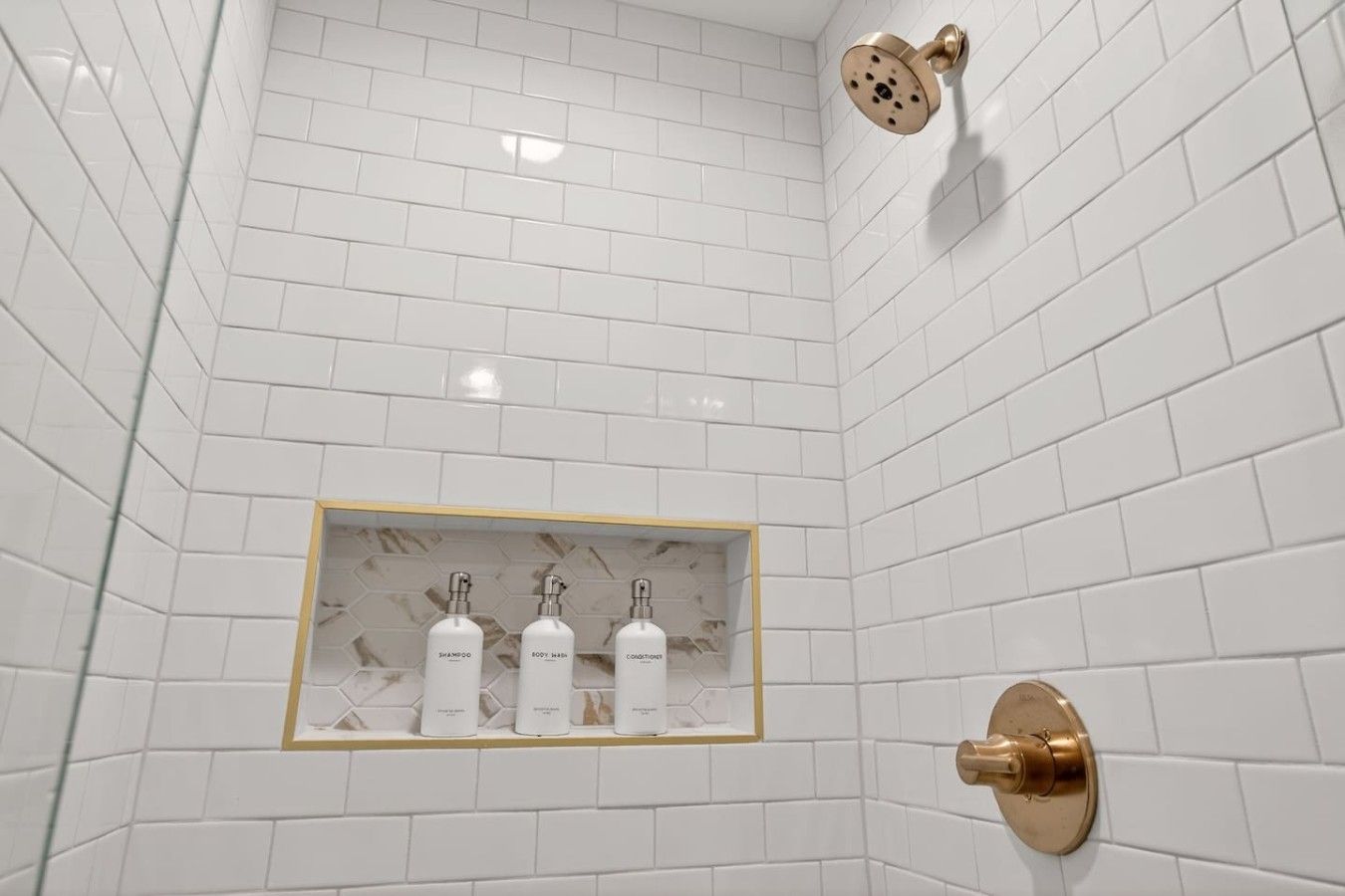 A bathroom with white subway tiles, a gold showerhead, and a shelf with three bottles of shampoo, body wash, and conditioner.