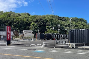 Een hotelentree met een traditioneel Japans gebouw, een omheind parkeerterrein en een uitzicht op een beboste heuvel.