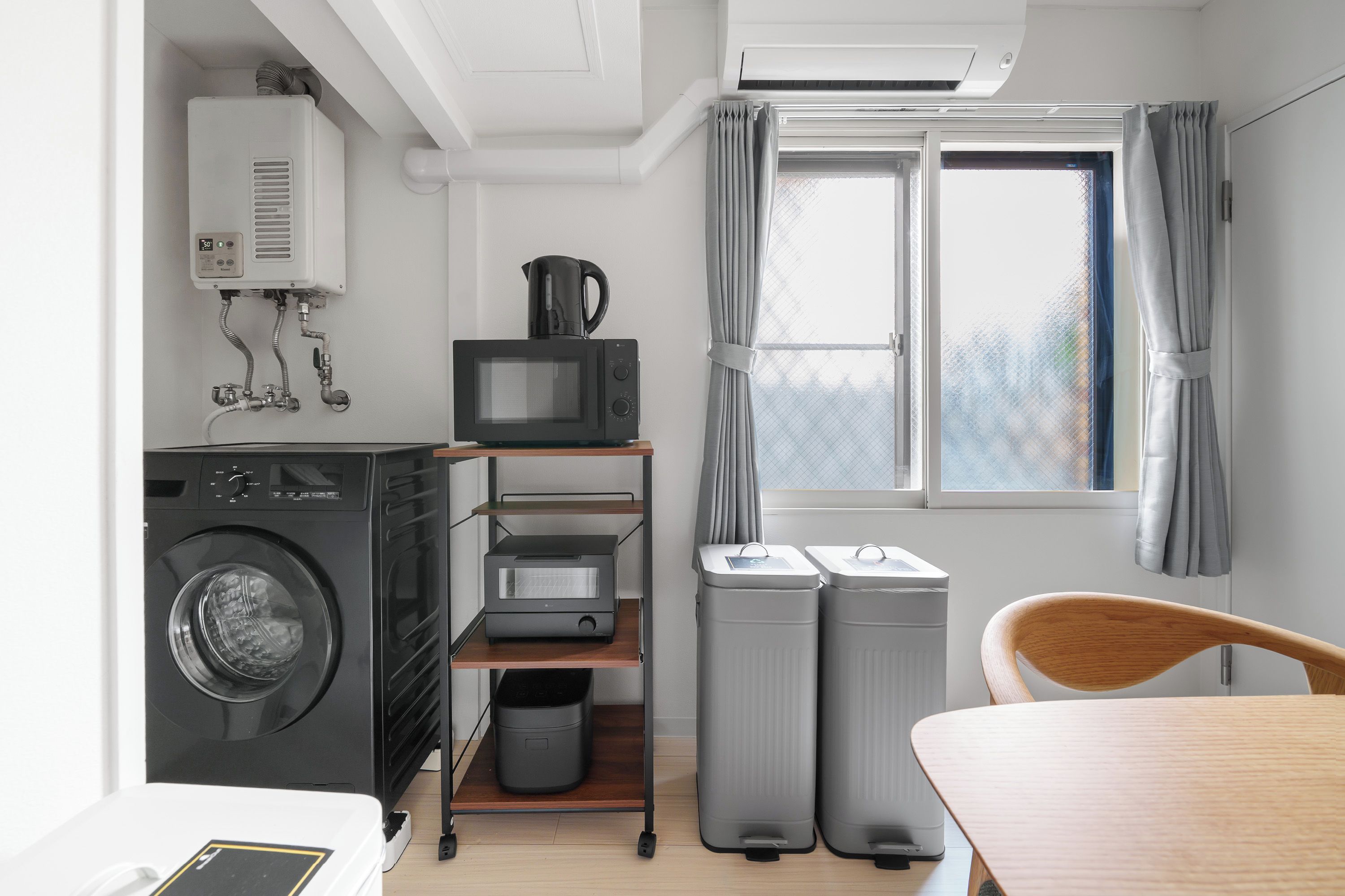 A compact hotel room with a washing machine, a microwave, a small kitchenette, a window with curtains, and a wooden chair.