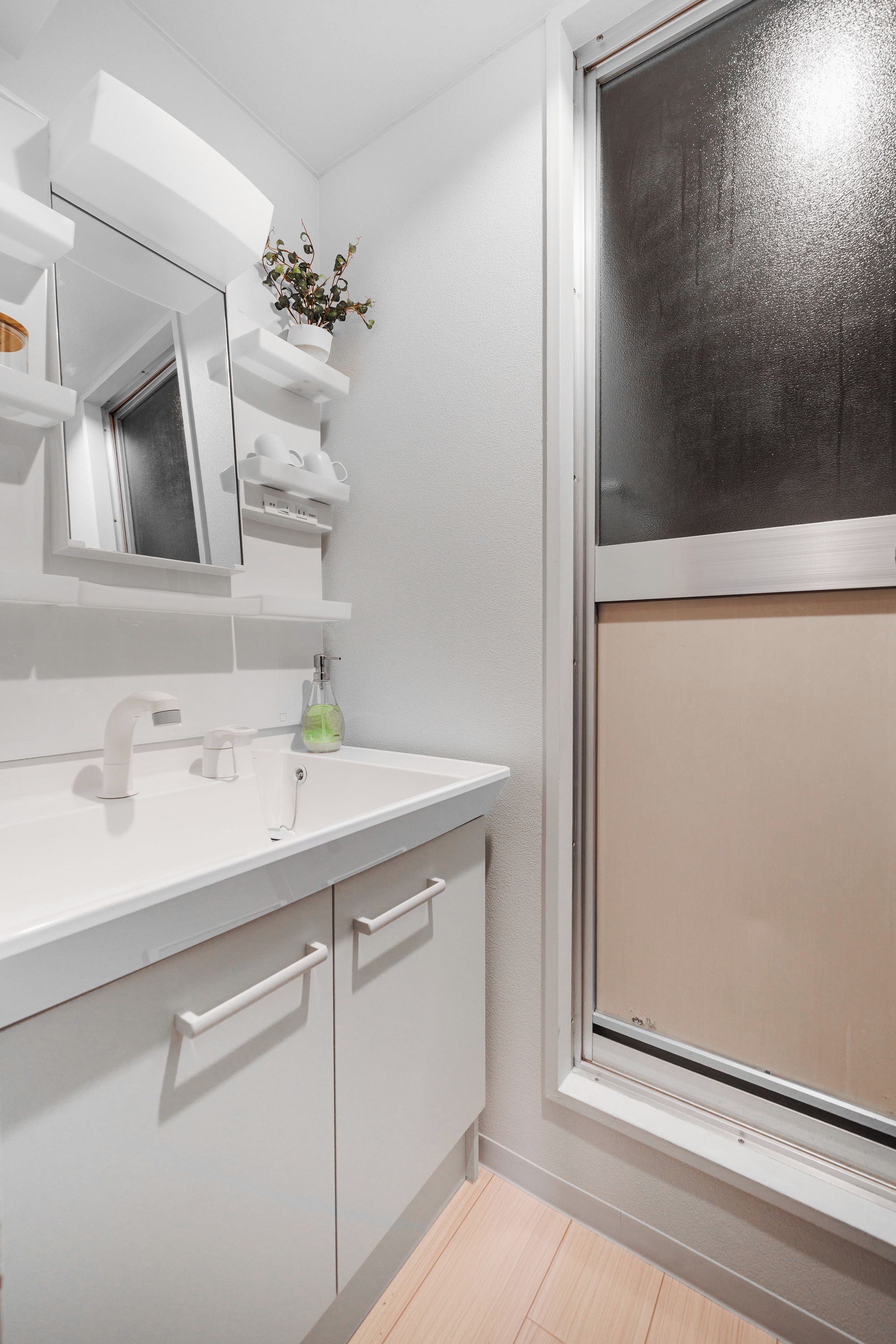 A modern bathroom with a white sink, mirror, and cabinets, a potted plant, and a window with a view.