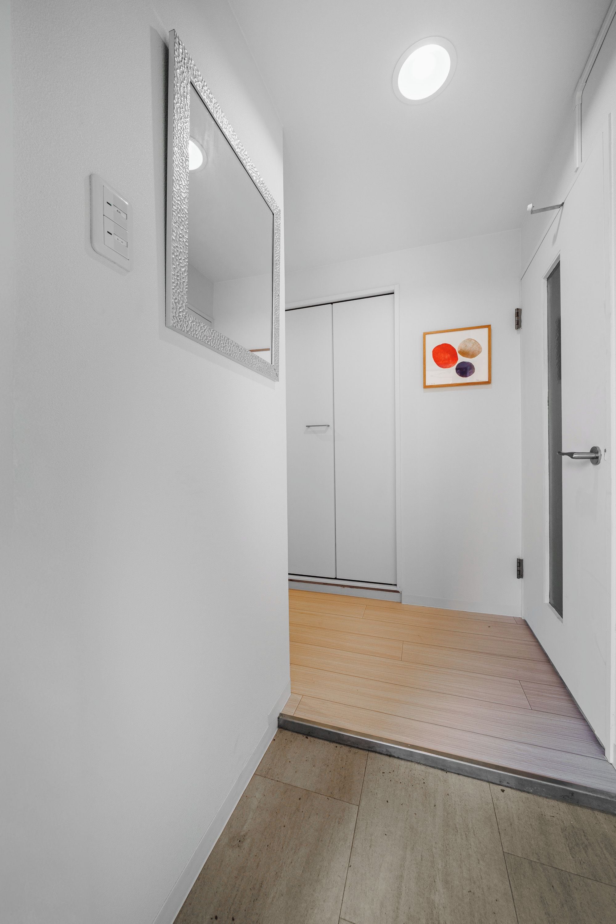 A hallway with a white wall, a mirror, a door, and a wooden floor.
