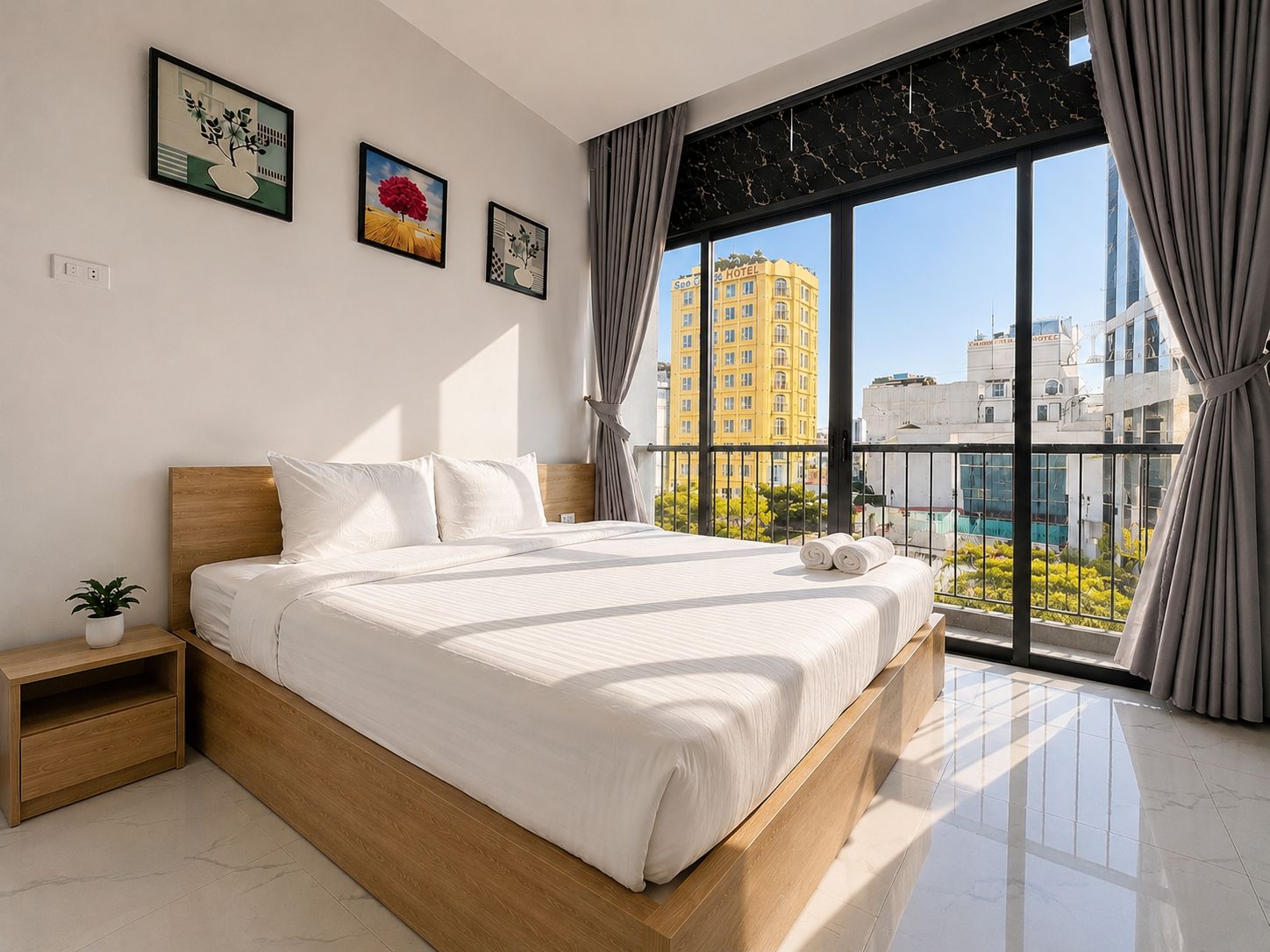 Ett modernt hotellrum med en stor säng, ett nattduksbord, en krukväxt och utsikt över staden genom stora fönster.