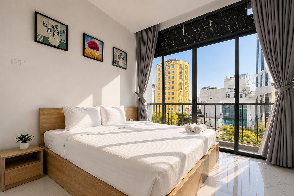 Quarto de hotel moderno com uma cama grande, uma mesa de cabeceira, um vaso de planta e vista da cidade através de amplas janelas.