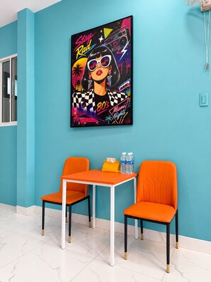 Quarto com paredes azul-turquesa, uma grande obra de arte vibrante com tema retrô, duas cadeiras laranja, uma mesa branca e um par de garrafas de água sobre a mesa.