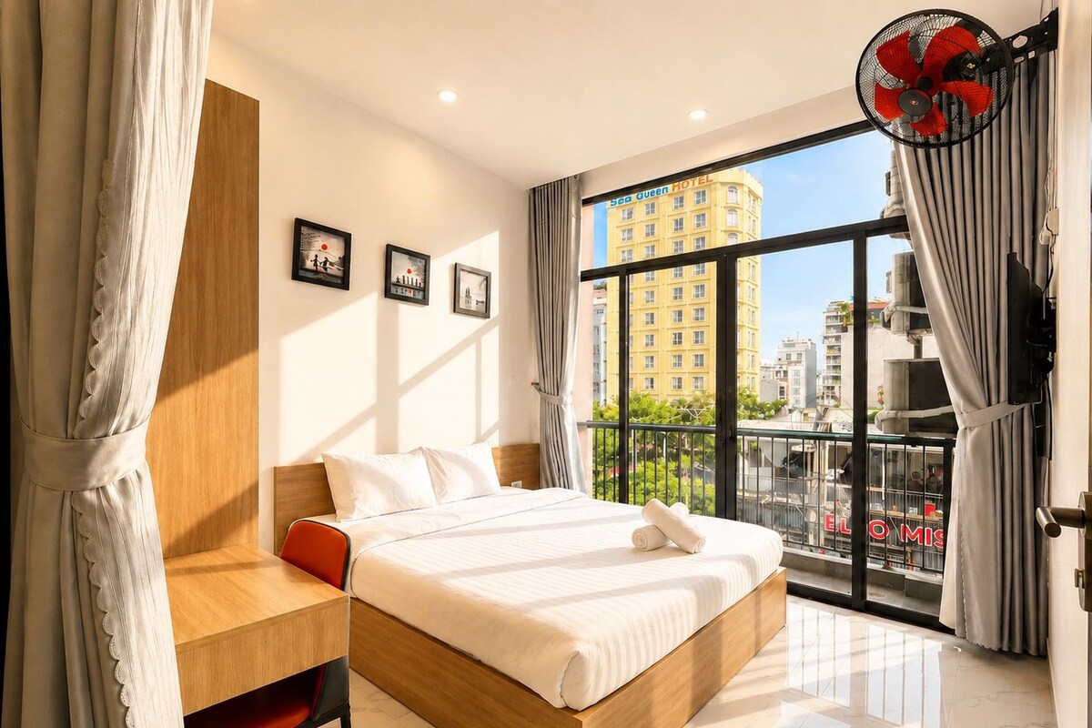 Quarto de hotel com cama, escrivaninha e uma grande janela com vista para a cidade.