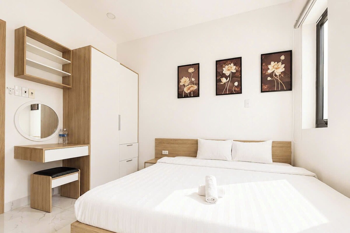 Quarto de hotel moderno com uma cama grande, um guarda-roupa de madeira, uma penteadeira com espelho e três quadros florais pendurados na parede.