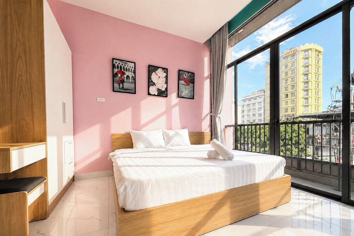 Quarto com cama, guarda-roupa e uma grande janela com vista para prédios.